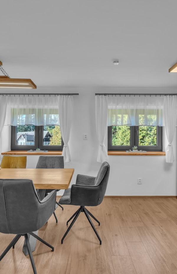 Modern dining room and kitchen with a wooden table, gray chairs, large windows with white curtains, wooden flooring, and a kitchen area with wooden cabinets and a gray refrigerator.