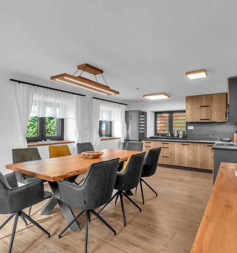 Modern dining and kitchen area with a wooden table, eight gray chairs, wooden flooring, and a kitchen with wooden cabinets and black countertops in the background.