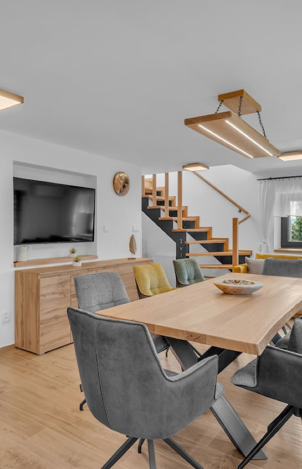 A modern living and dining area with a wooden table, gray chairs, a yellow sofa, a flat-screen TV on the wall, and a staircase leading upstairs.