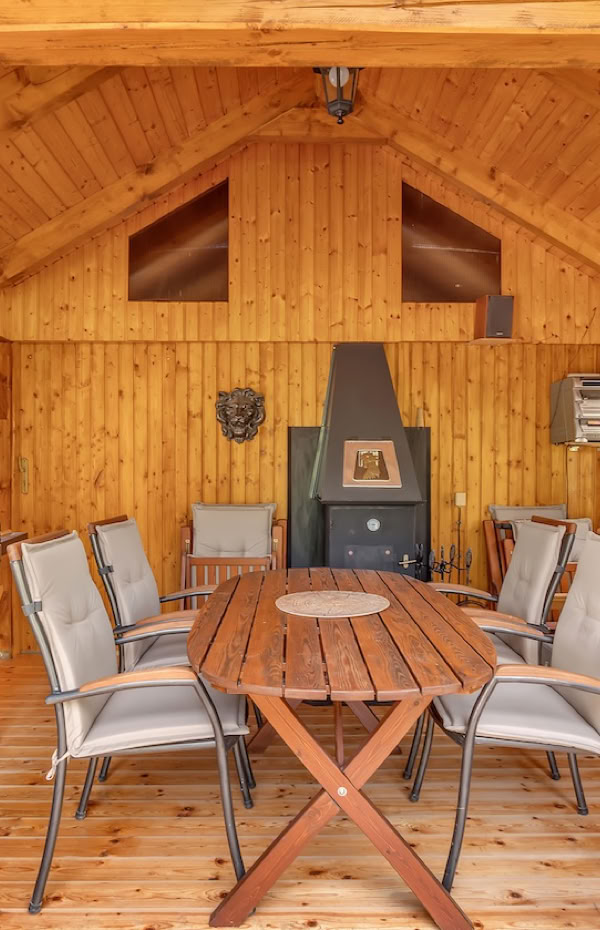 Wooden interior of a cabin with a long table and chairs, a sofa, a fireplace, and a barbecue grill.