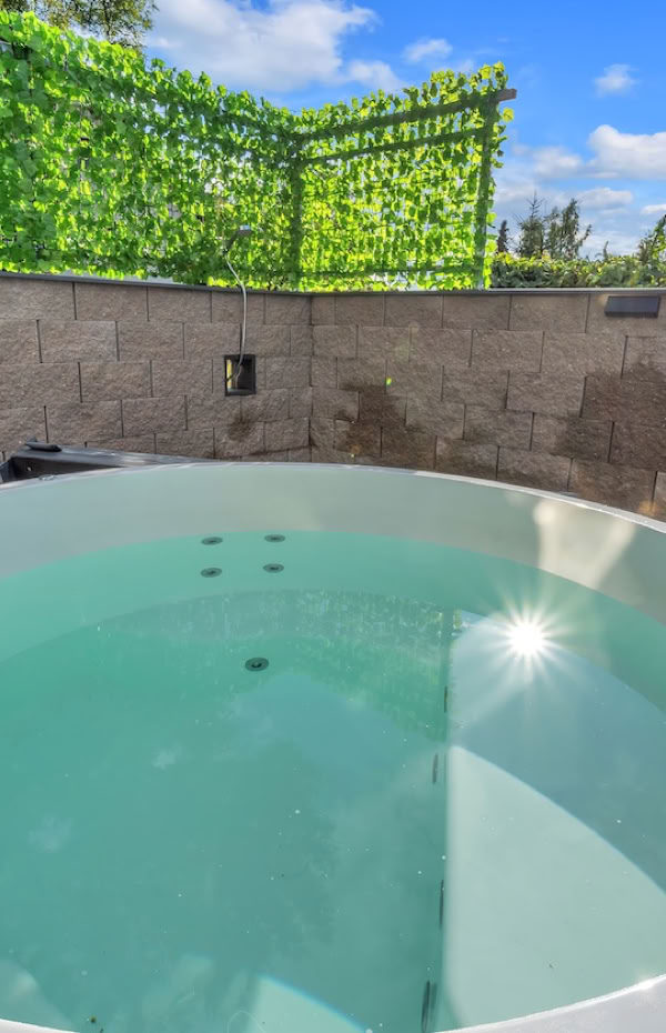 Round outdoor hot tub filled with water, surrounded by a stone wall and greenery, with a chimney-like structure on the right.