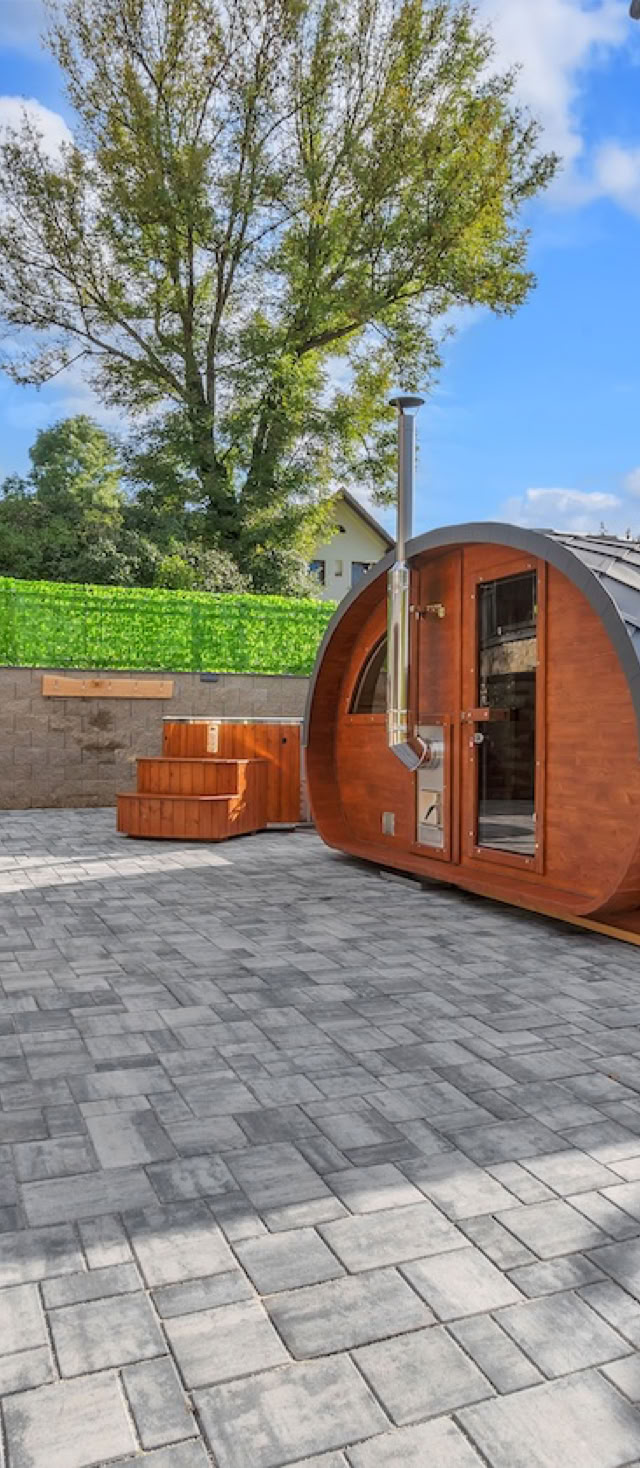 A courtyard with a wooden pod structure, two orange deck chairs, stone walls, and a tree in the background under a blue sky.
