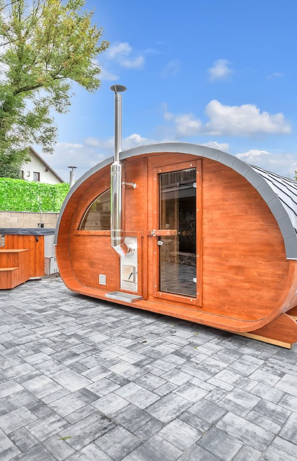 A wooden barrel-shaped sauna with a chimney on a paved patio, surrounded by a stone wall and greenery under a blue sky.