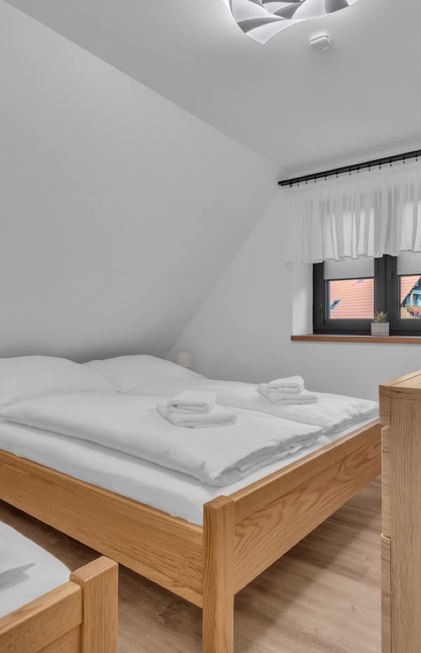 A bedroom with two single beds, a wooden dresser with a television on top, a window with white curtains, and a small bedside table with folded towels.